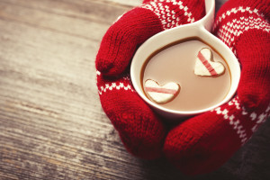 Hands in mittens holding hot cup of coffee with marshmallow hearts on wooden background